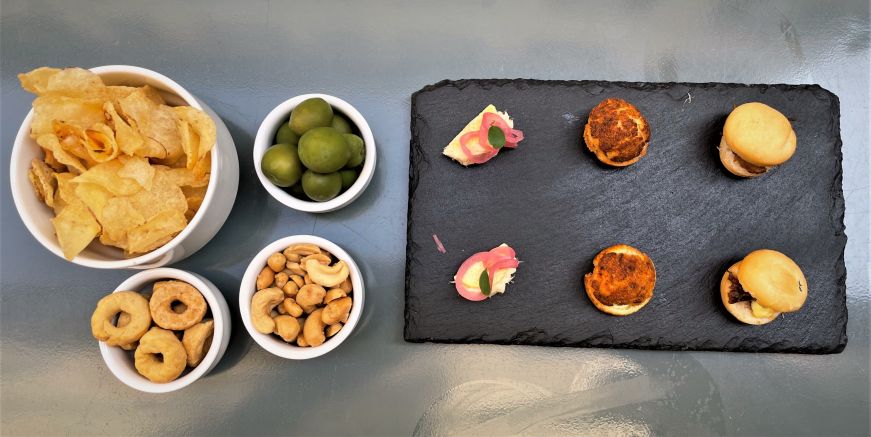 Top down view of table with small bowls of olives, nuts, crackers, and potato chips and a plate of miniature sandwiches