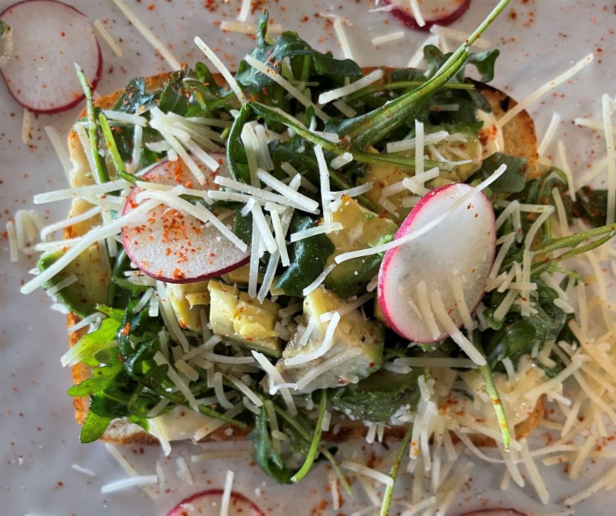 Slice of toast topped with arugula, avocado chunks, radishes, and shredded Parmesan