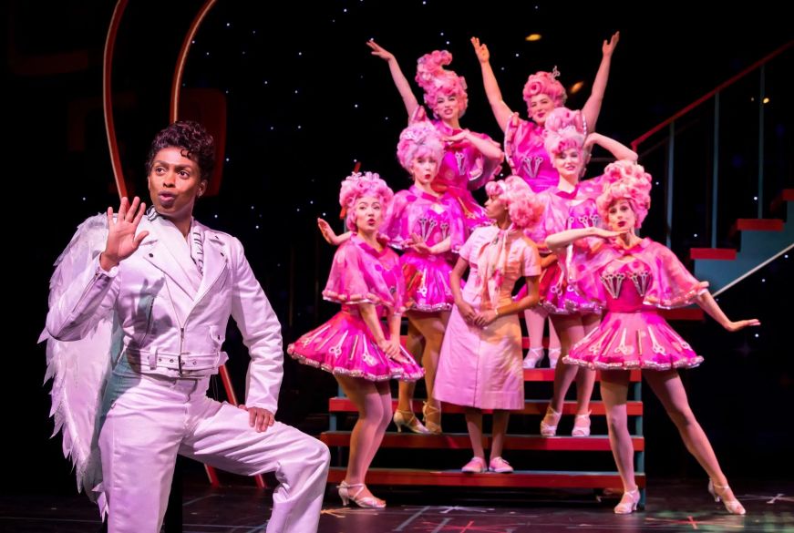 Man dressed as an angel singing on stage with pink-clad dancers in the background