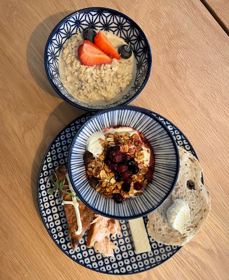 Top down view of a bowl of muesli and plate with a yogurt parfait, bread, smoked salmon, and cheese