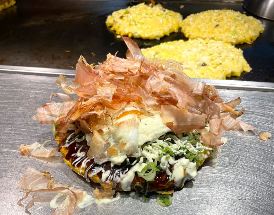 Okonomiyaki piled with bonito with more okonomiyaki cooking on a griddle in the background