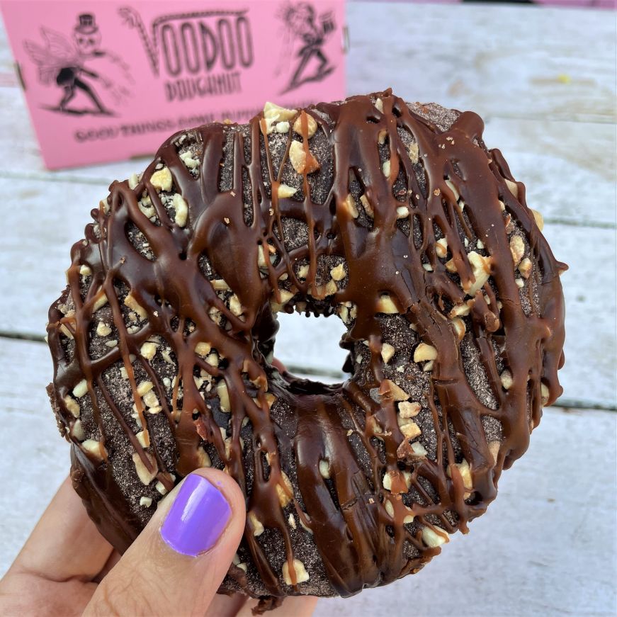 Stacy's hand holding a raised doughnut topped with chocolate frosting, hot chocolate powder, peanuts, and chocolate drizzle