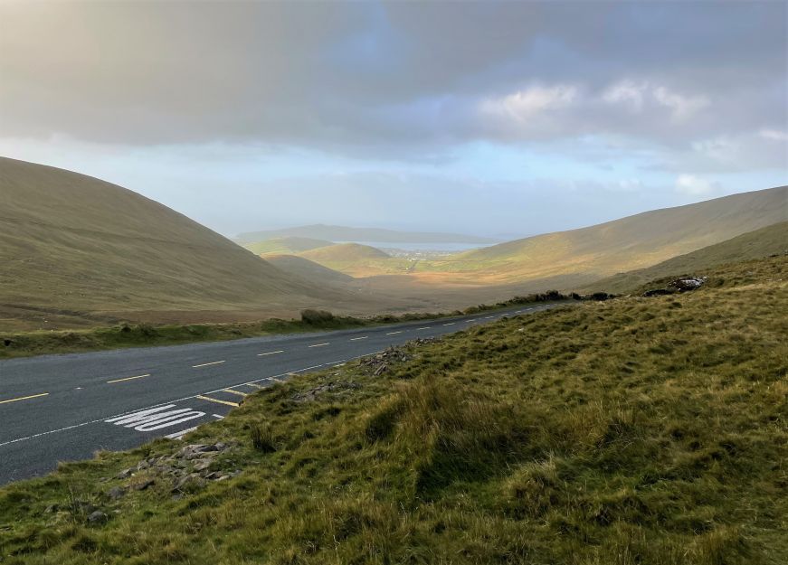 Narrow two-lane blacktop road with rolling hills, small town, and ocean in background