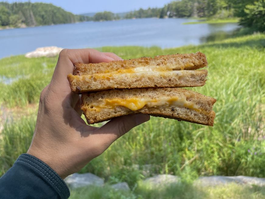 Grilled cheese on multigrain bread, White Sails Bakery & Deli Hand holding a grilled cheese sandwich with a body of water in the background
