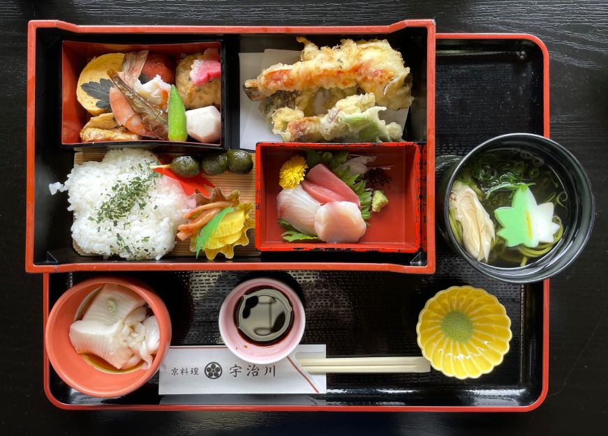 Bento box with compartments filled with rice, sashimi, pickles, tempura, and other items