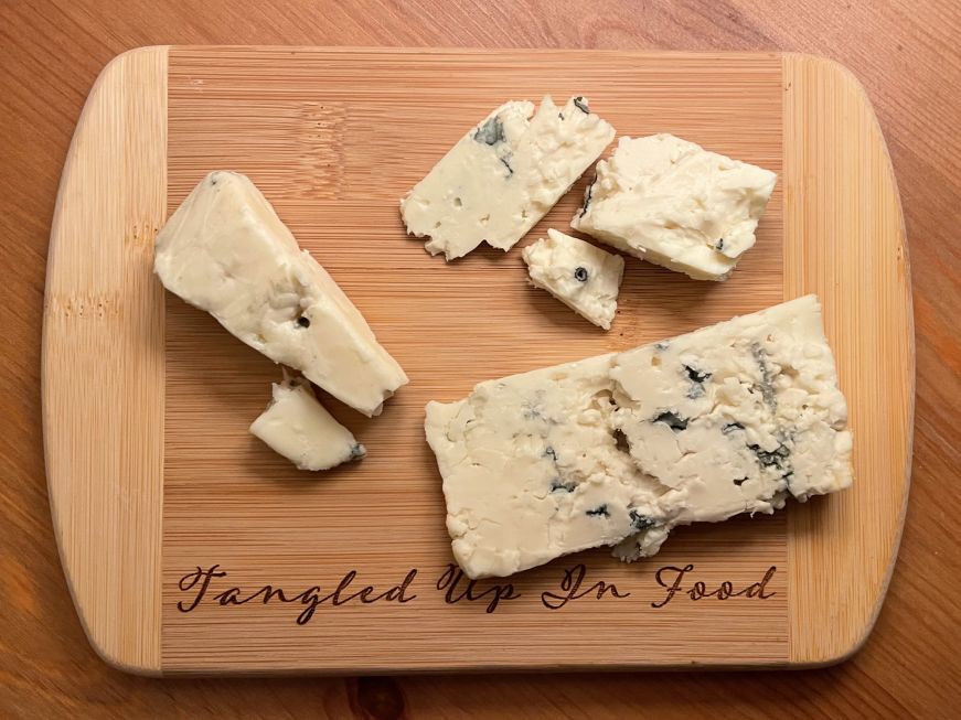 Several pieces of blue cheese on a cutting board with a Tangled Up In Food logo